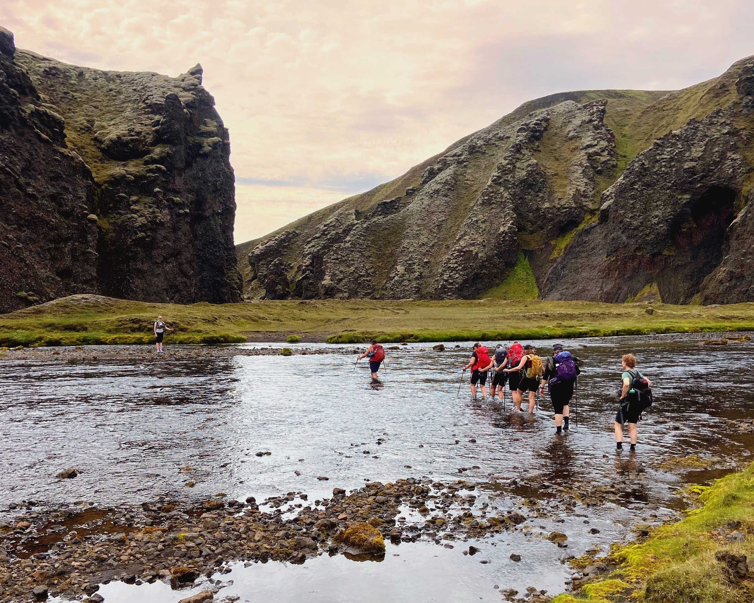 A small group wades a shallow river through a green highland canyon under a soft sky